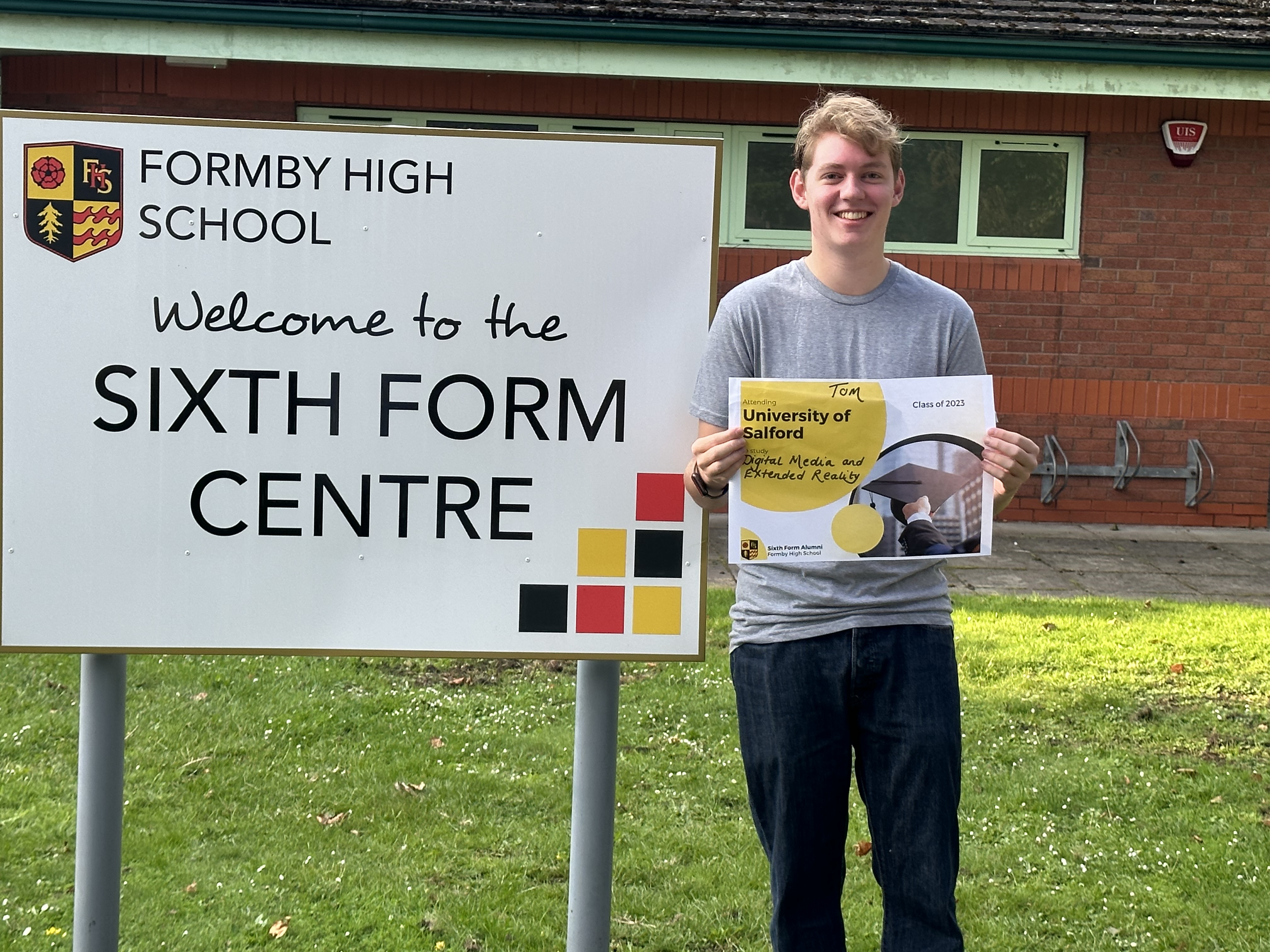 Thomas infornt of Formby High School Sixthform holding a sign that says Tom Attending the University of Salford to study Digital Media and Extended Reality