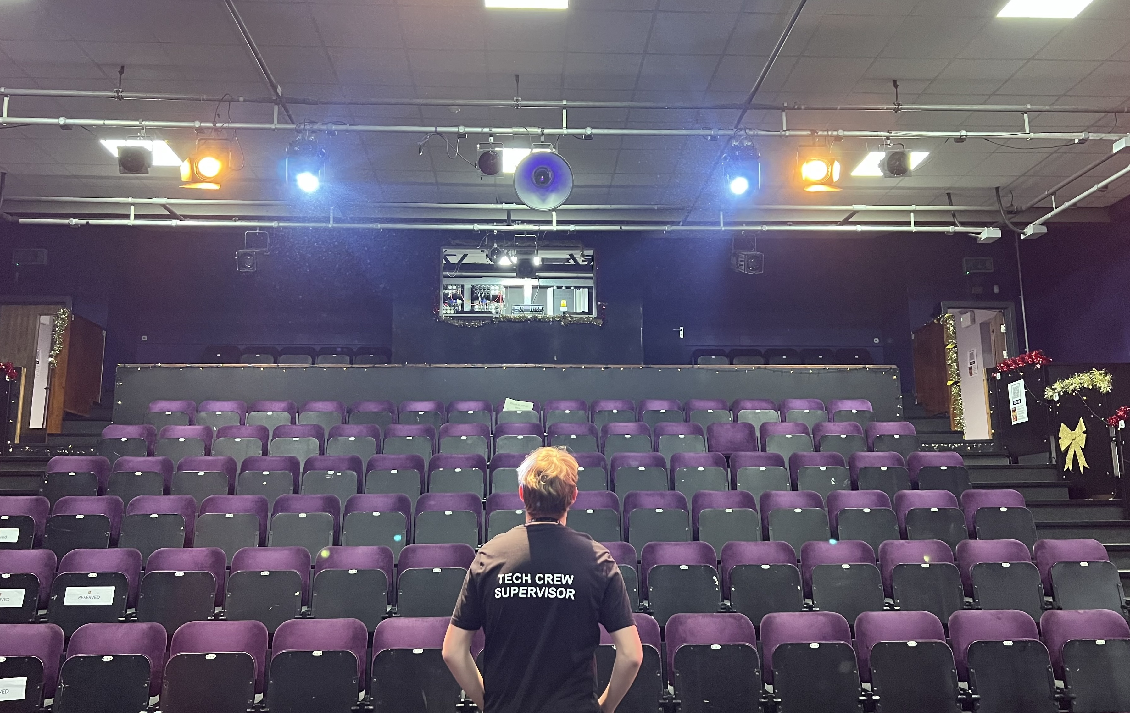 Thomas stood in a theatre with his back to the camera with a shirt that says Tech Crew Tech Suporvisor
